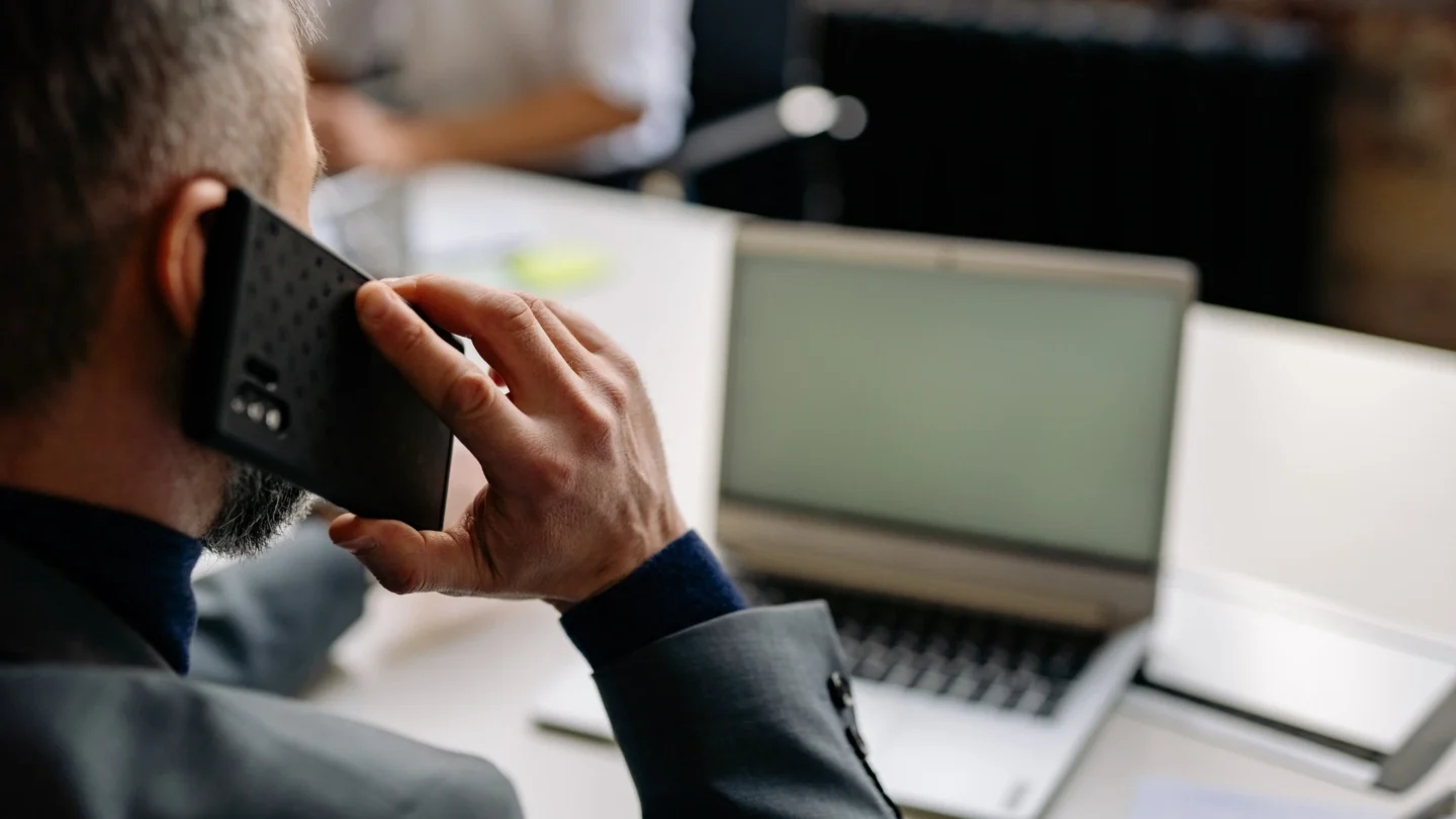 Man holding a phone, obviously calling, sitting behind a laptop