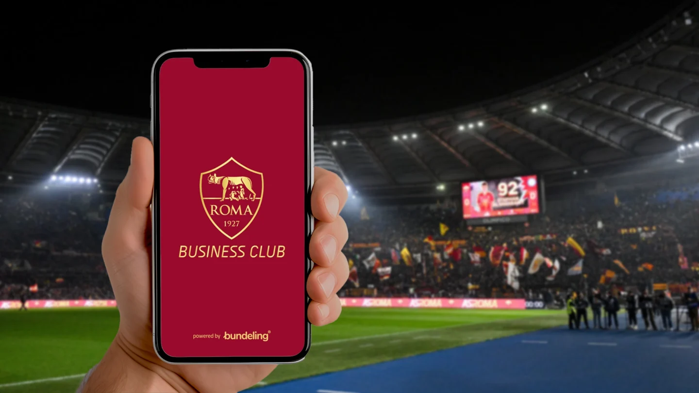 A hand holding the AS Roma Bundeling app with the AS Roma stadium in the background
