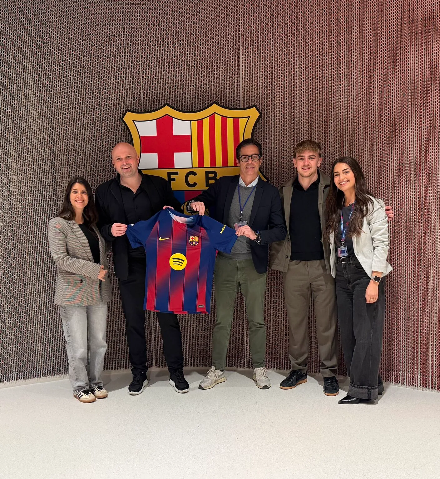 3 men and to women smiling towards the camera in front of the FC Barcelona logo, holding a FC Barcelona shirt. 